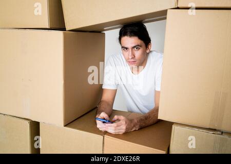 Young man holding smart phone while trapped under cardboard containers Stock Photo