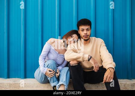 Girlfriend leaning on boyfriend's shoulder while sitting against blue wall Stock Photo