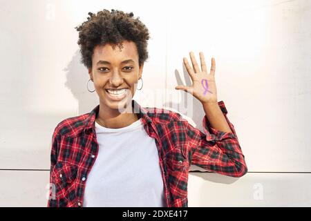 Cheerful young woman with purple awareness symbol on palm against white wall during Womens Day Stock Photo