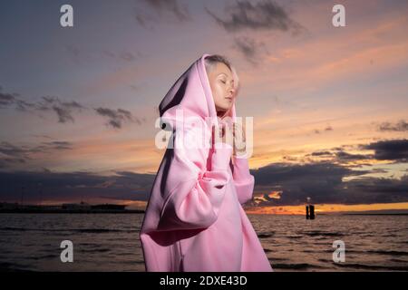 Young woman wearing pink hooded shirt standing on beach at sunset Stock Photo