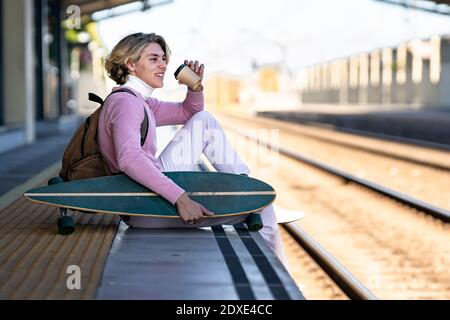 Smiling young man holding reusable cup while sitting with longboard at edge of railroad platform Stock Photo