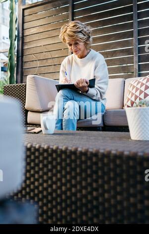 Mature woman writing in diary sitting on sofa at rooftop garden Stock Photo
