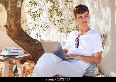 Thoughtful young man with laptop looking away while sitting in backyard Stock Photo