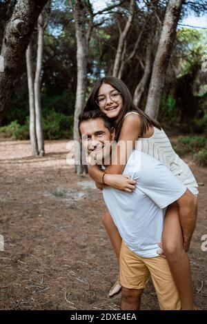 Happy father giving piggyback ride to daughter in forest during vacation Stock Photo