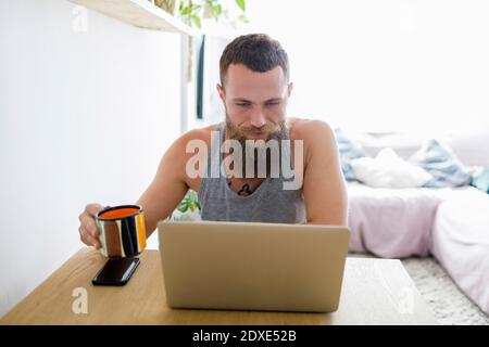 Bearded man using laptop while holding coffee cup at home Stock Photo