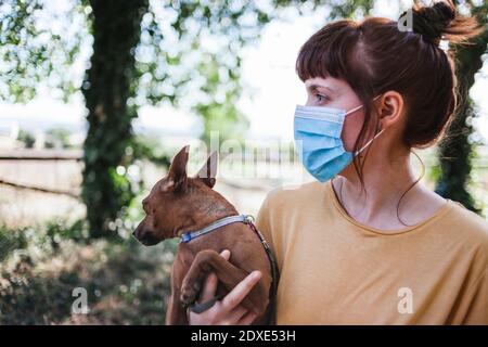 Young woman wearing face mask carrying dog while walking at park during COVID-19 Stock Photo