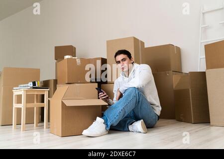 Young man using mobile phone while sitting against cardboard boxes in new house Stock Photo