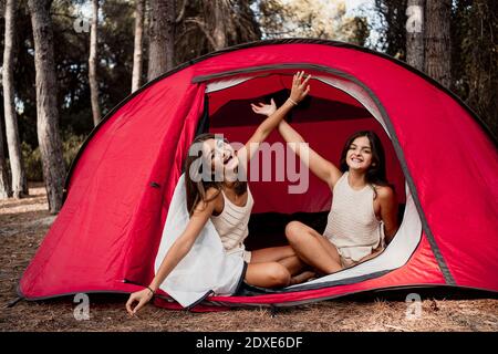 Cheerful sisters with hands raised sitting in tent at campsite during vacation Stock Photo