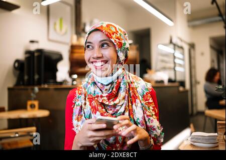 Smiling Muslim woman in floral hijab looking away while holding mobile phone at cafe Stock Photo