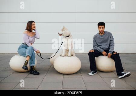 Couple sitting with social distance by dog on white concrete ball against wall Stock Photo