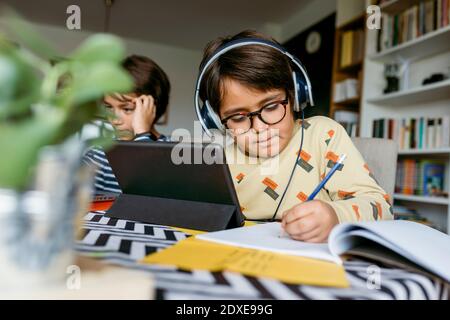 Boy writing notes through digital tablet sitting with male friend during e-Learning at home Stock Photo