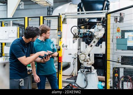 Male coworkers holding digital tablet looking at robotic arm while standing in manufacturing industry Stock Photo