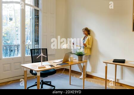 Businesswoman examining blueprint while standing against wall in office Stock Photo