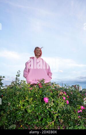 Young woman with pink hair wearing pink hooded shirt near rose bush Stock Photo