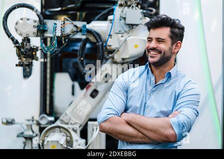 Smiling male entrepreneur with arms crossed looking away while standing by robotic arm at industry Stock Photo