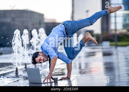 Businessman using laptop while doing handstand on footpath by fountain Stock Photo