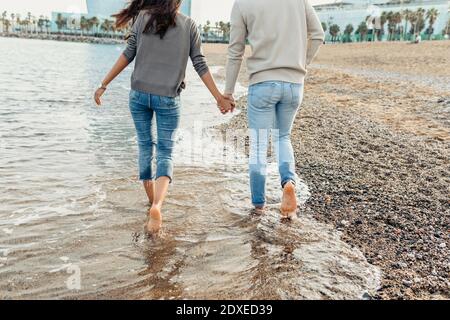 Young couple holding hands while walking on water at beach Stock Photo
