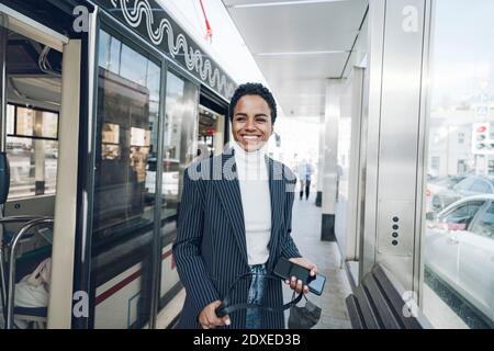 Happy businesswoman with mobile phone at bus stop during COVID-19 outbreak Stock Photo