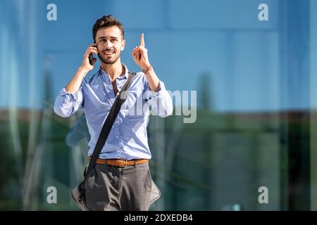 Businessman wearing crossbody bag pointing upward while talking on mobile phone outdoors Stock Photo
