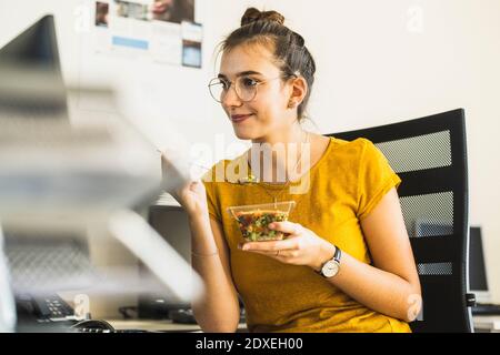 Smiling businesswoman eating food while looking at computer sitting at office Stock Photo
