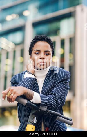 Confident young businesswoman leaning on handlebar of electric push scooter Stock Photo