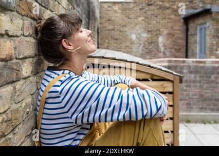 Smiling woman leaning on wall while sitting at back yard Stock Photo