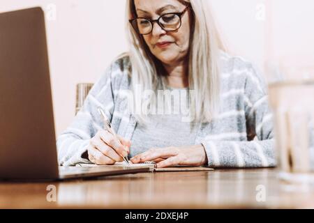 Senior woman writing notes during online consultation at home Stock Photo