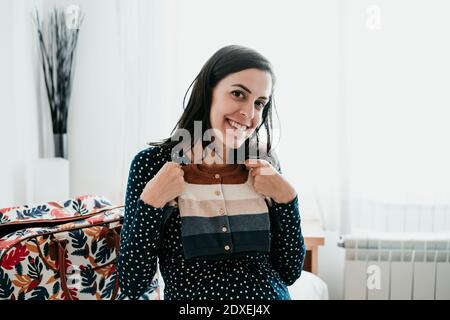 Happy pregnant woman holding baby clothes at home Stock Photo