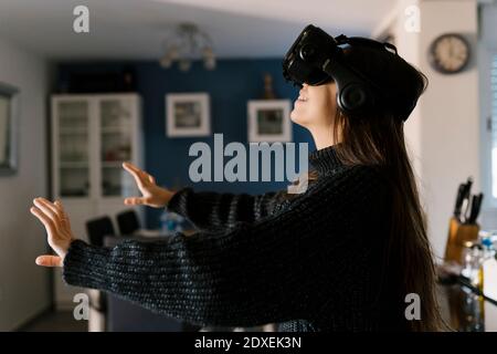 Playful teenage girl wearing virtual reality simulator eyeglasses sitting at home Stock Photo