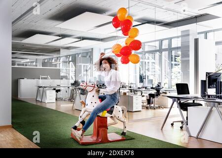 Playful businesswoman holding balloons while sitting on rocking horse at work place Stock Photo