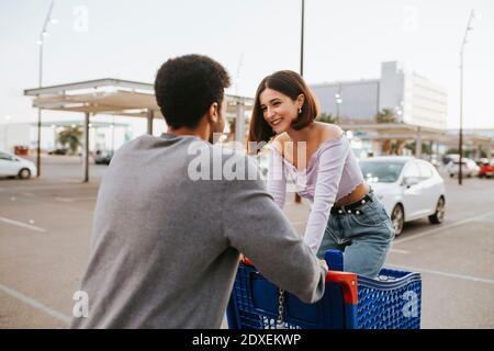 Young man pushing woman standing in shopping cart on road Stock Photo