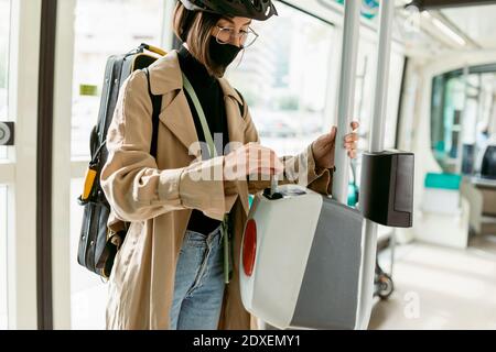 Woman wearing face mask and cycling helmet putting ticket in ticket validation machine while standing in tram Stock Photo