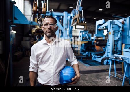 Male entrepreneur wearing protective eyewear while holding helmet standing in factory Stock Photo