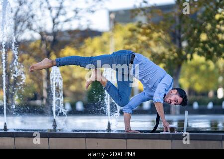 Young businessman using laptop while doing handstand on retaining wall Stock Photo