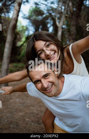 Father giving piggyback ride to daughter with arms outstretched in forest during vacation Stock Photo