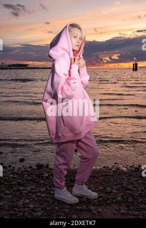 Portrait of young woman wearing pink hooded shirt standing on beach at sunset Stock Photo