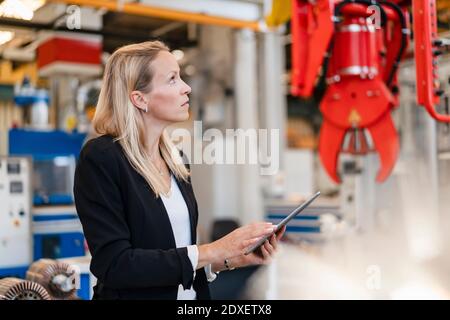 Blond female entrepreneur analyzing claw while holding digital tablet standing in factory Stock Photo
