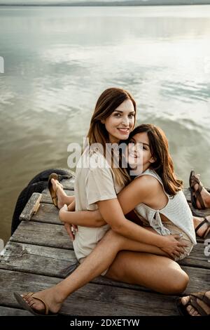 Mother and daughter embracing each other while sitting at jetty against lake Stock Photo