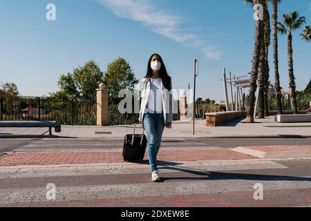 Woman with luggage crossing street against blue sky on sunny day during pandemic Stock Photo