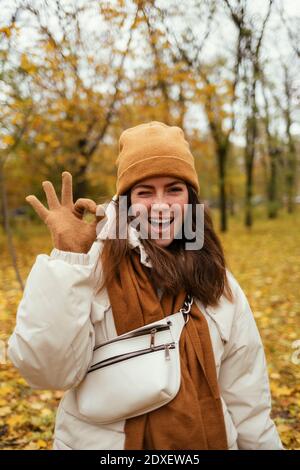 Happy young woman winking while making OK sign in autumn park