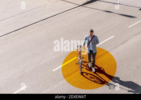 Man with wheeling bicycle using mobile phone while walking on road with gold colored circle Stock Photo