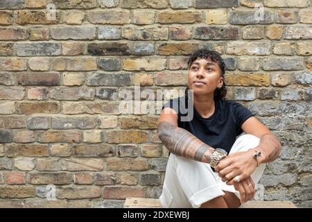 Thoughtful woman looking away while sitting against brick wall at back yard Stock Photo