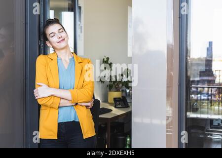 Young businesswoman with arms crossed and eyes closed standing by door at office