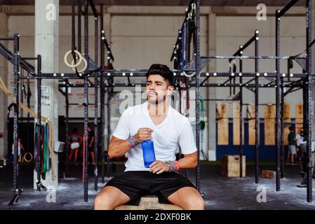 Young man holding water bottle while sitting on box at gym Stock Photo