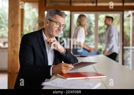 Businessman using digital tablet while standing by table with colleagues in background at home Stock Photo