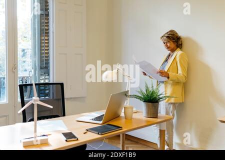 Female entrepreneur analyzing blueprint while leaning against wall in office Stock Photo