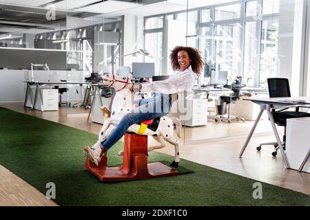 Happy businesswoman on rocking horse pretending to ride in office Stock Photo