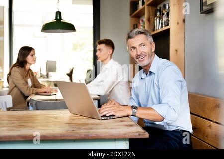 Smiling businessman using laptop while sitting with colleagues in background at office Stock Photo