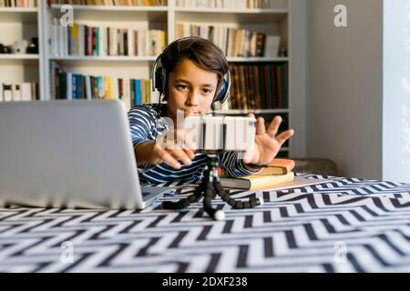 Boy adjusting smart phone on tripod sitting by laptop at table Stock Photo