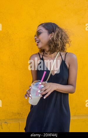 Happy young woman holding smoothie against yellow wall Stock Photo